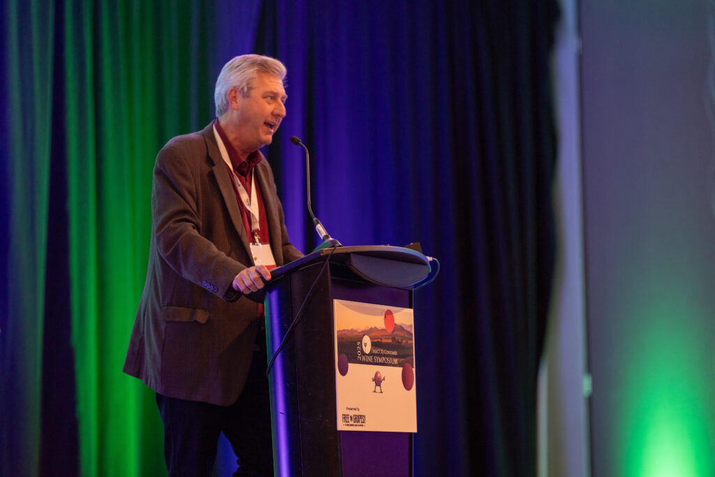 A man speaks at a podium on stage with dark curtains and colorful lighting in the background.