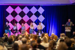 Five speakers sit on stage with a moderator at a conference, addressing an audience in a large room.