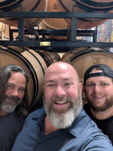 Three men smiling in front of stacked wine barrels, showcasing the rich grape culture in a winery or cellar setting.