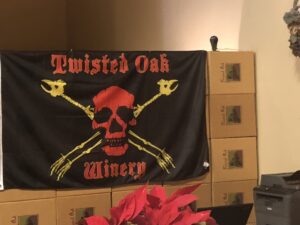 A Twisted Oak Winery flag with a red skull and yellow bones stands atop boxes; a red plant grows in the foreground.